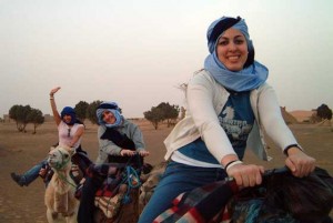 Three female SJSU students riding camels in desert. 