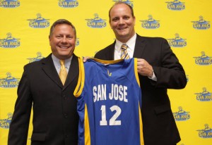 Two white men wearing suites, on left is Tim La Kose. Tim is holding up a SJSU basketball Jersey with the number 12 on it. They are standing infront of a yellow background with SJSU spartan logos.