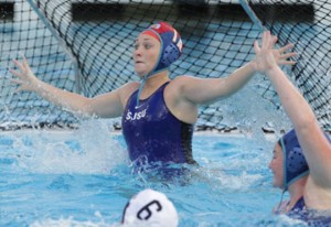 Lauren Lewis in the pool attempting to block the opposing team from scoring a goal, wearing a blue swimsuite.