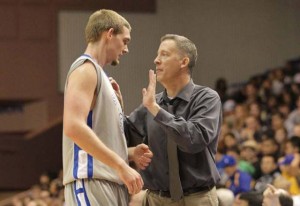 George Nessman, wearing a suite, talks to a basketball player during a game who is wearing a grey jersey with blue stripes. 