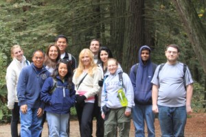 Students gather in front of sequoia trees to pose for a picture at the Nicene Marks State Park. They are wearing jackets and have backpacks on ready to hike.