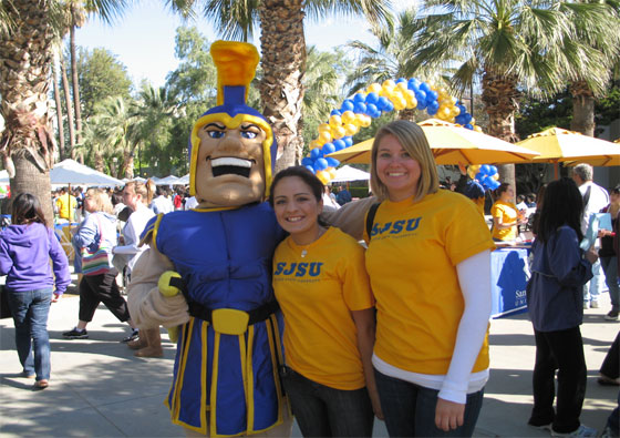 Two female students in gold SJSU shirt posing with Sammy Spartan.