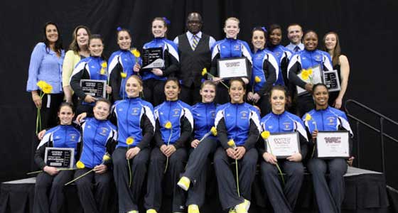 Gymnastics team poses in uniform holding their awards with their coach. (black background.)