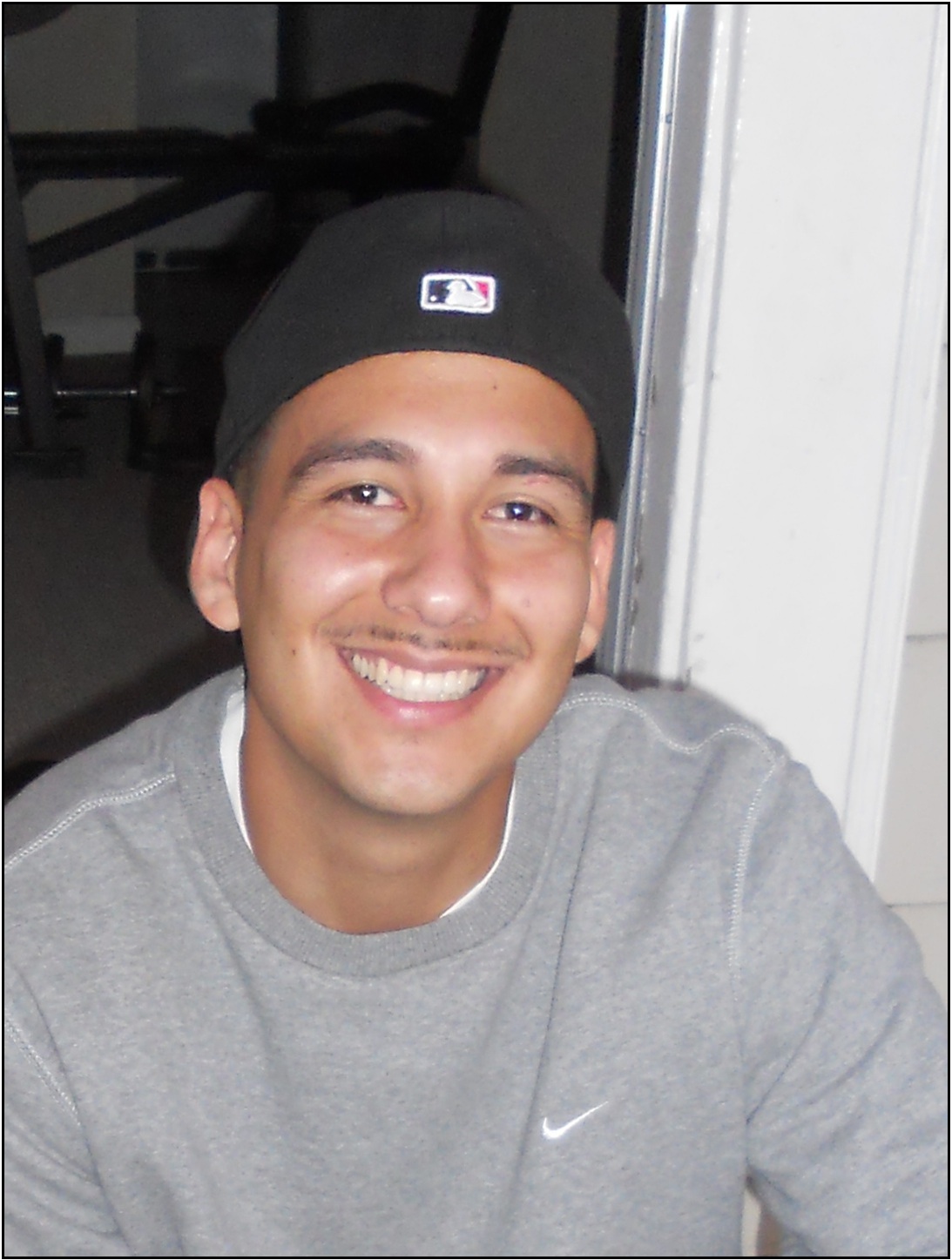 Daneil Guzman is standing in front of a doorframe, smiling to the camera wearing a baseball cap and sweatshirt