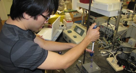 male researcher holding a syringe-like instruments over a plate in a lab full of equipment