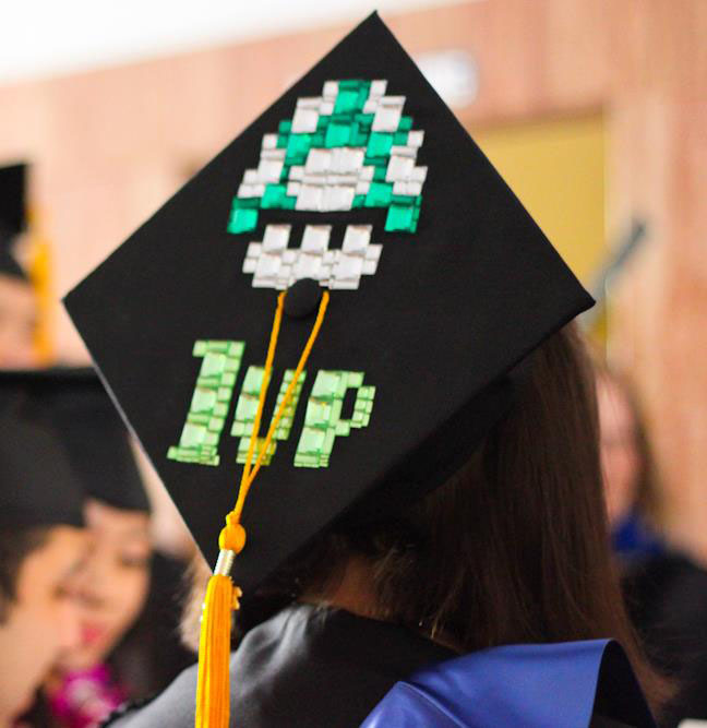 Close-up photo of a graduation cap with yellow tassel that has a green-and-white toadstool with the phrase 1up from the Mario video games. Photo by Christina Olivas