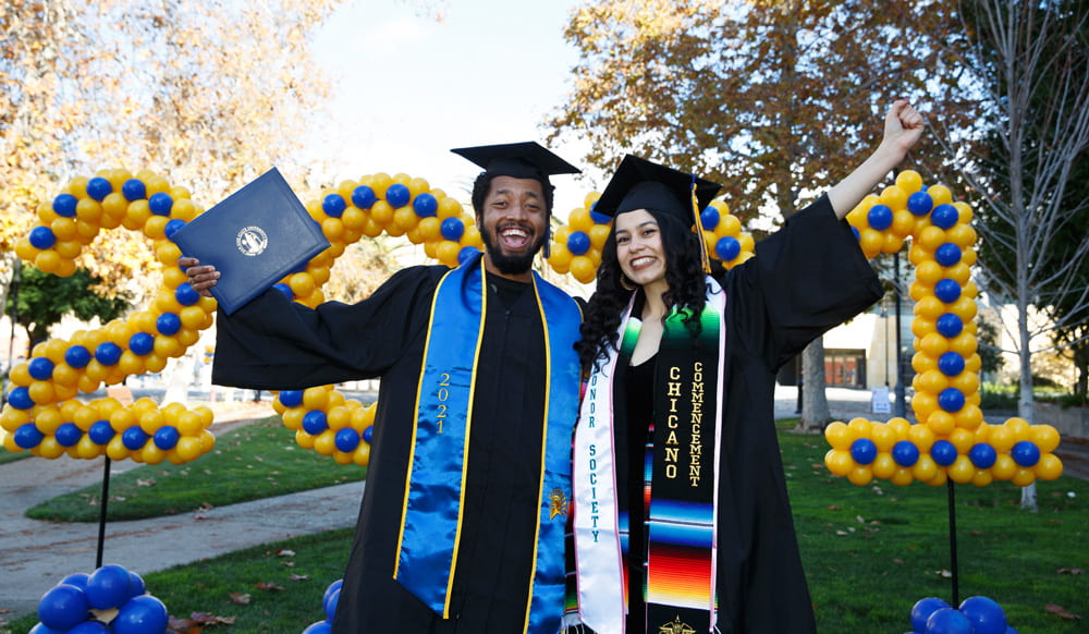 Two students in regalia pose in front of 2021 balloons.