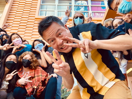 Professor David Chai leaning forward holding up the peace sign with both hands with a group of people behind him also throwing up the peace sign.