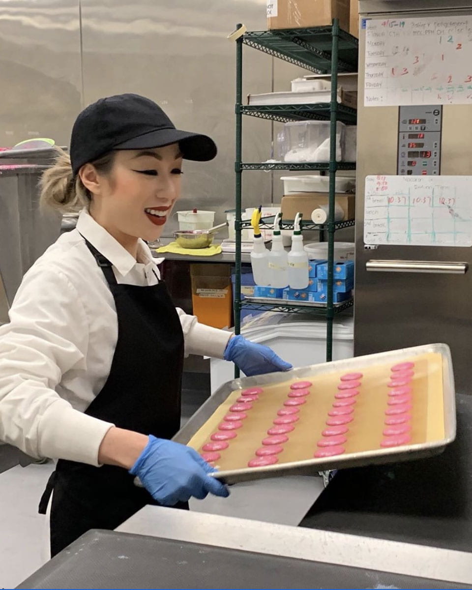 Gwen Nguyen in an apron holding a tray of bright pink macarons that are ready to be put in the oven to bake.